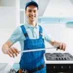 Smiling male furniture maker in uniform holds measuring tape in the kitchen. Handyman installing garniture, repairing service at home